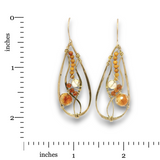 A pair of gold teardrop dangle earrings with amber and glass bead details, shown next to a black measurement ruler on a white background.