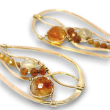 A pair of gold-toned statement earrings with organic, flowing wire designs, featuring round and faceted amber-orange and clear gemstones on a white background.