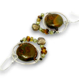 A pair of handmade dangle earrings featuring polished brown and amber stone beads, small green and brown accent beads, and silver wire wrapping on white hooks, shown against a plain white background.