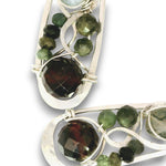 Silver-toned statement earrings featuring faceted green tourmaline and dark brown to black tourmaline stones, set in an open, swirling metal design on a white background.