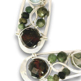 Silver-toned statement earrings featuring faceted green tourmaline and dark brown to black tourmaline stones, set in an open, swirling metal design on a white background.