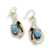 A pair of gold teardrop dangle earrings featuring faceted blue teardrop gemstones, small dark round beads, and dark round accent stones on a white background.