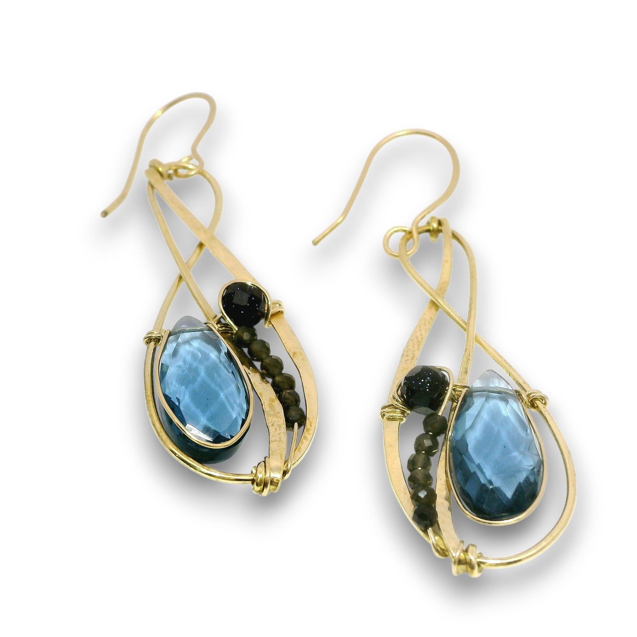 A pair of gold teardrop dangle earrings featuring faceted blue teardrop gemstones, small dark round beads, and dark round accent stones on a white background.