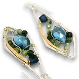 A pair of handmade dangle earrings featuring faceted blue and dark blue teardrop-shaped gemstones, surrounded by small green and gray beads, set in gold and silver-tone metal wire frames.