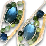 Close-up of two matching earrings with large faceted blue gemstones, green and blue beads, and gold and silver metal settings on a white background.