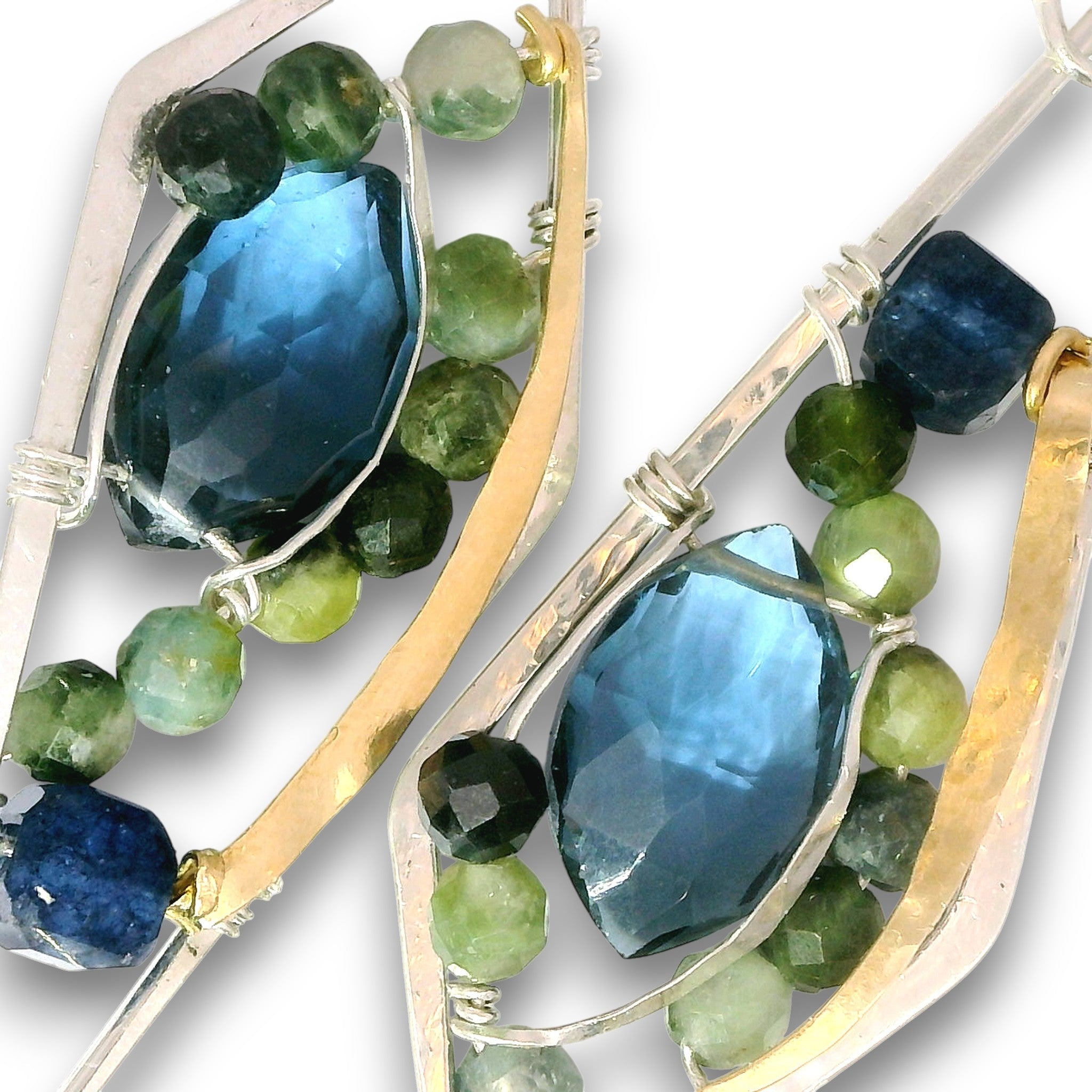 Close-up of two matching earrings with large faceted blue gemstones, green and blue beads, and gold and silver metal settings on a white background.