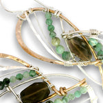 Close-up of handmade wire-wrapped earrings featuring green and smoky brown gemstone beads, with silver and gold-toned metal wires on a white background.