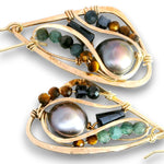 A pair of gold-toned teardrop-shaped earrings featuring a large iridescent pearl, faceted green and brown beads, and dark rectangular gemstones on a white background.