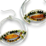 A pair of silver hoop earrings with oval bezel settings holding a polished amber-orange center stone, surrounded by rows of small green and brown beads, on a white background.