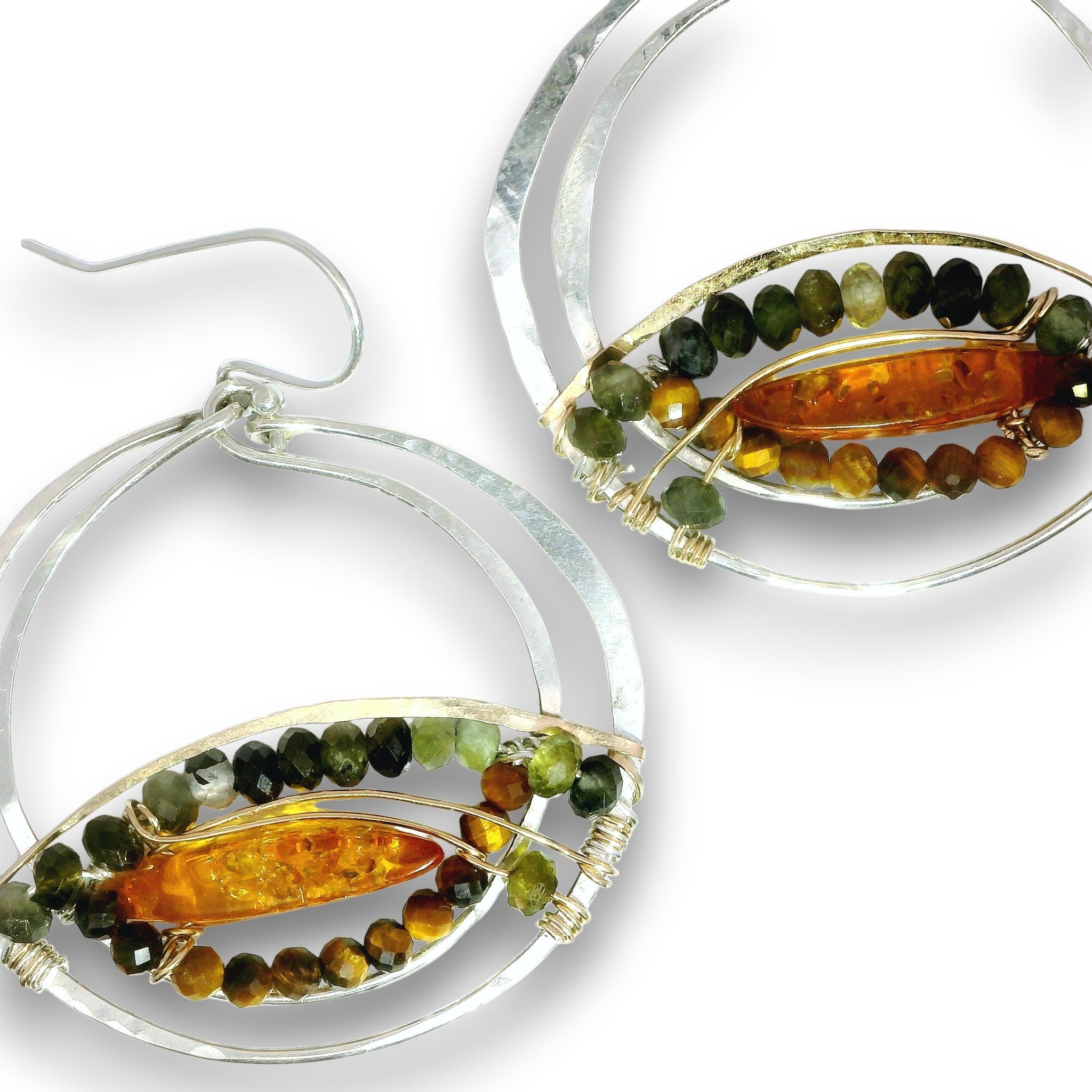 A pair of silver hoop earrings with oval bezel settings holding a polished amber-orange center stone, surrounded by rows of small green and brown beads, on a white background.