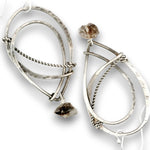 A pair of silver-toned geometric dangle earrings with hammered and twisted wire details, each featuring a small rough brown stone at the top and a larger polished stone at the bottom, shown on a white background.