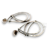 A pair of modern, abstract dangle earrings made of hammered and twisted silver-tone metal, each featuring a small, rough, clear faceted gemstone, shown on a white background.