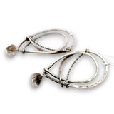 A pair of modern, abstract dangle earrings made of hammered and twisted silver-tone metal, each featuring a small, rough, clear faceted gemstone, shown on a white background.