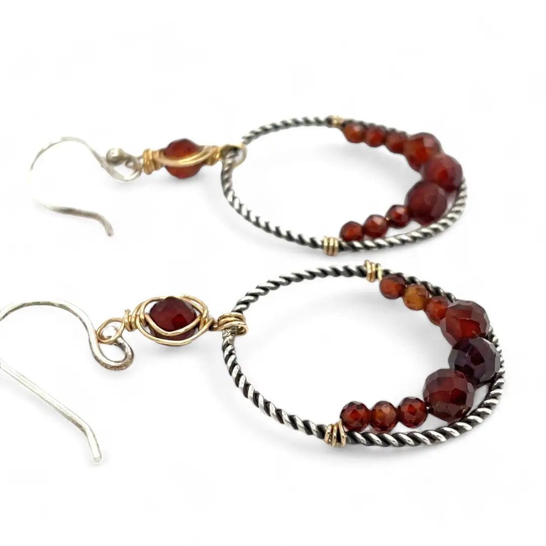 A pair of twisted oxidized sterling silver hoop earrings with garnet gemstone accents and faceted hessonite garnets on gold filled wire