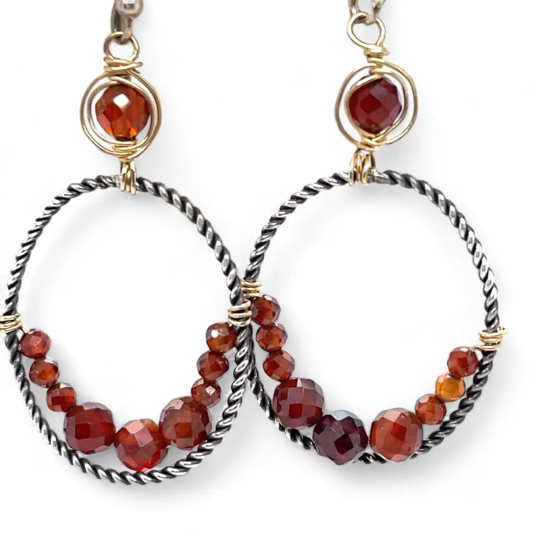 A pair of twisted oxidized sterling silver hoop earrings with garnet gemstone accents and faceted hessonite garnets on gold filled wire