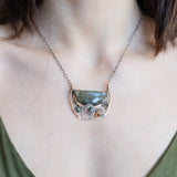 A flashy green labradorite half moon is set inside a brass cup pendant encrusted with oxidized sterling silver coils with pyrite, green kyanite, and citrine gemstone, hanging from a textured darkened sterling silver chain necklace