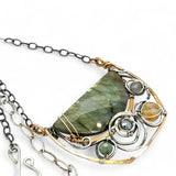 A flashy green labradorite half moon is set inside a brass cup pendant encrusted with oxidized sterling silver coils with pyrite, green kyanite, and citrine gemstone, hanging from a textured darkened sterling silver chain necklace