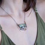 A flashy green labradorite half moon is set inside a brass cup pendant encrusted with oxidized sterling silver coils with pyrite, green kyanite, and citrine gemstone, hanging from a textured darkened sterling silver chain necklace