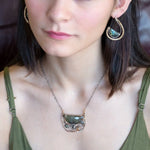 A flashy green labradorite half moon is set inside a brass cup pendant encrusted with oxidized sterling silver coils with pyrite, green kyanite, and citrine gemstone, hanging from a textured darkened sterling silver chain necklace