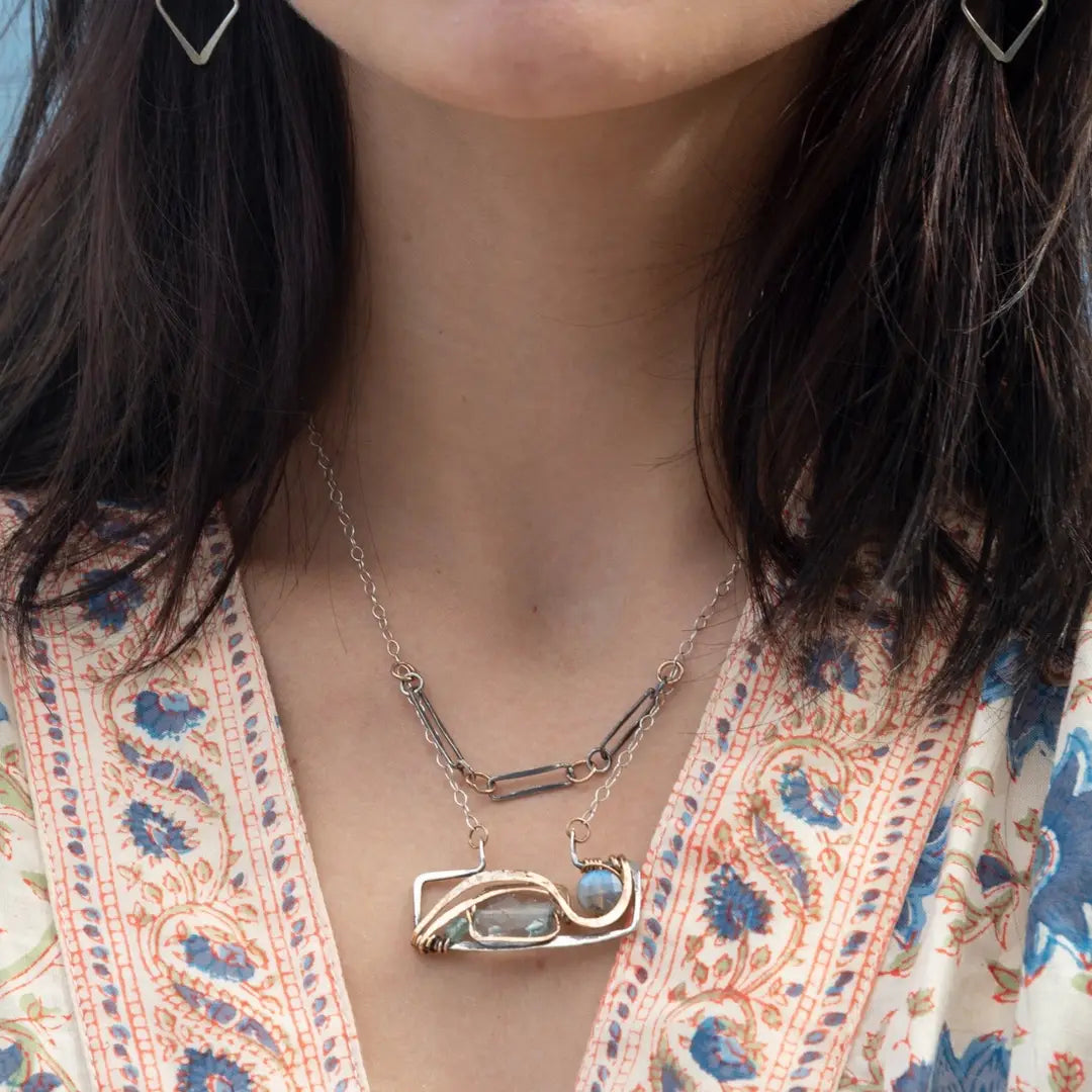 A double-layered necklace with a geometric design, featuring a rectangular darkened sterling silver pendant with a fluorite gemstone, flashy labradorite gemstone, and bronze accents, connected to chain links and additional bronze rings.