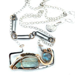A double-layered necklace with a geometric design, featuring a rectangular darkened sterling silver pendant with a fluorite gemstone, flashy labradorite gemstone, and bronze accents, connected to chain links and additional bronze rings.