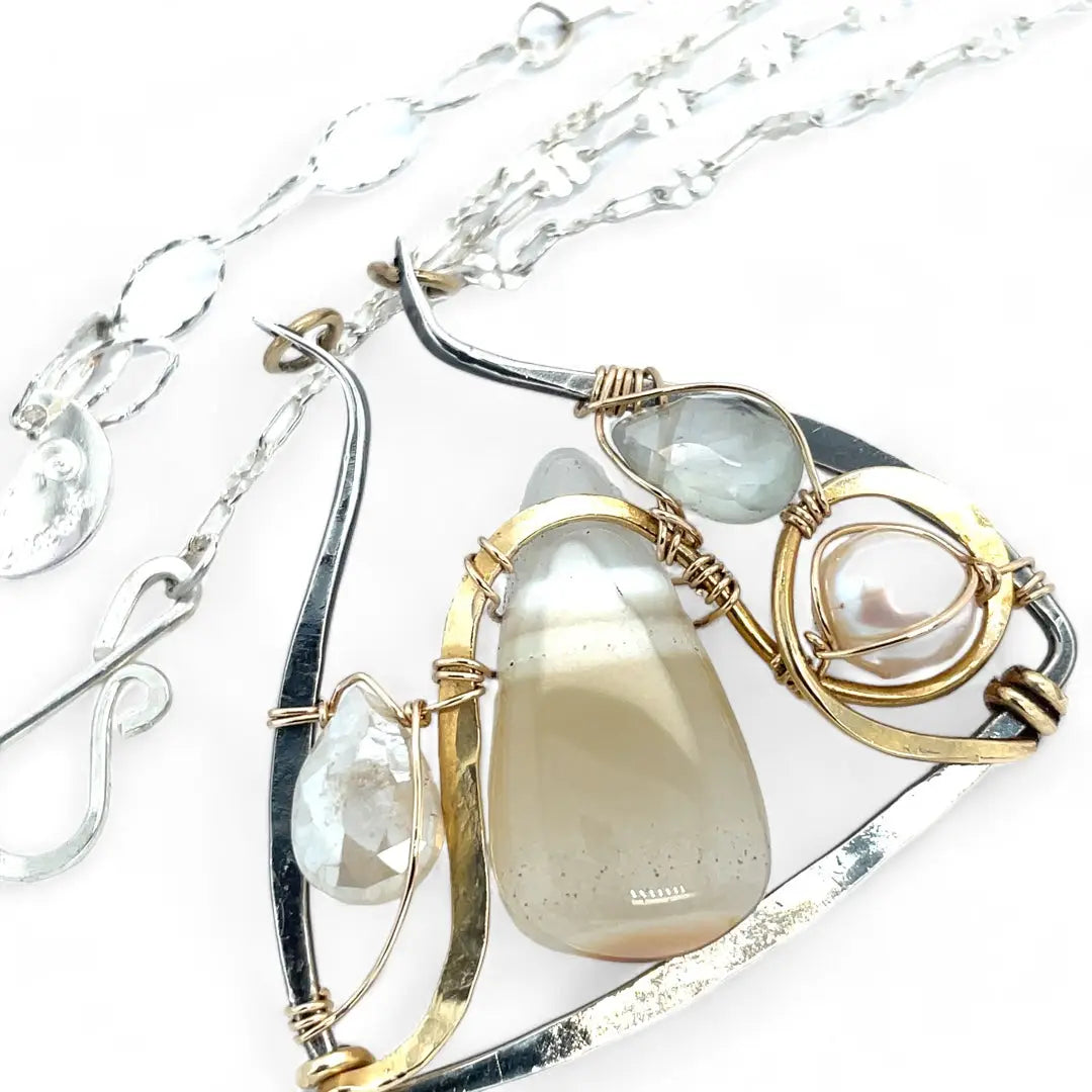 A darkened sterling silver moth-shaped pendant featuring a smooth cream agate gemstone, white sapphire, and jade drops, complemented by a fresh water pearl, with layers of gold filled and hammered brass wires