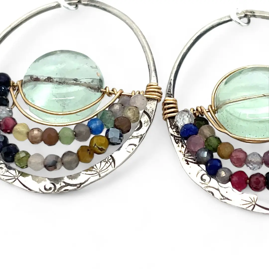 A pair of sterling silver hoop earringsthat have been stamped with dandelions and swirls, featuring a green fluorite smooth coin wrapped in gold filled wire and surrounded by a array of multicolored gemstones.