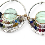 A pair of sterling silver hoop earringsthat have been stamped with dandelions and swirls, featuring a green fluorite smooth coin wrapped in gold filled wire and surrounded by a array of multicolored gemstones.
