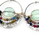 A pair of sterling silver hoop earringsthat have been stamped with dandelions and swirls, featuring a green fluorite smooth coin wrapped in gold filled wire and surrounded by a array of multicolored gemstones.