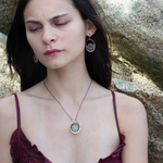 A pair of sterling silver hoop earringsthat have been stamped with dandelions and swirls, featuring a green fluorite smooth coin wrapped in gold filled wire and surrounded by a array of multicolored gemstones.