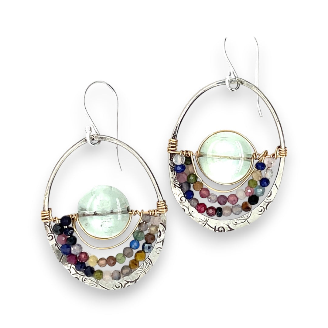 A pair of sterling silver hoop earringsthat have been stamped with dandelions and swirls, featuring a green fluorite smooth coin wrapped in gold filled wire and surrounded by a array of multicolored gemstones.