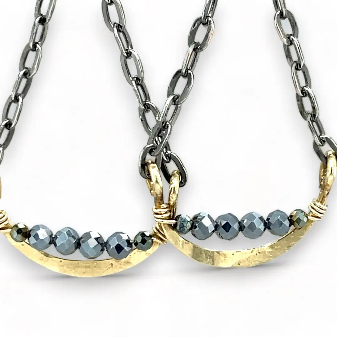 A pair of earrings featuring hand-formed brass arches with faceted pyrite gemstones, suspended from black chain links with sterling silver French hooks.
