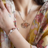 A darkened sterling silver pallete pendant necklace with bronze hammered swirls, featuring a series of faceted gemstones including amethyst and rhodocrosite, peach fresh water coin pearl, and crystal, complemented by a rose gold chain and a silver textured chain.