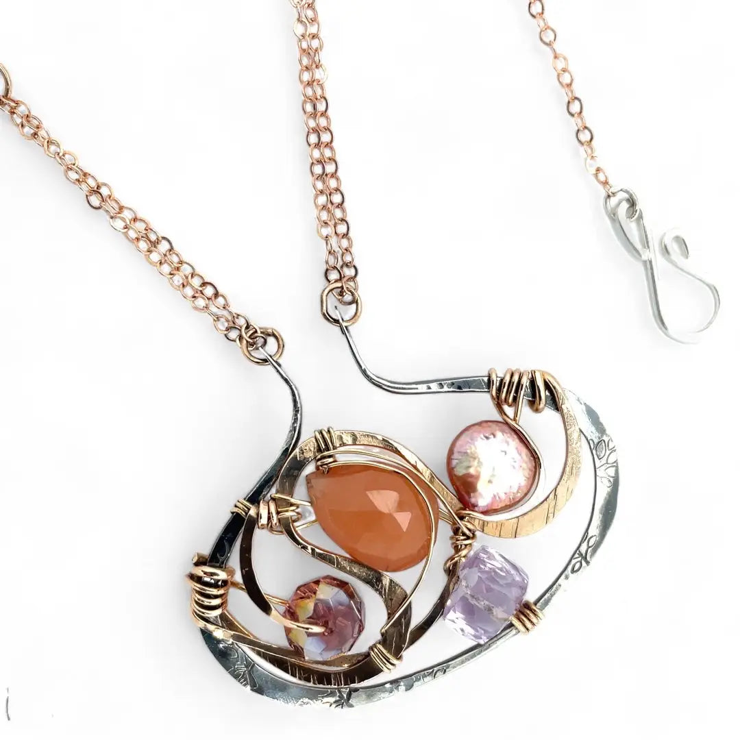 A darkened sterling silver pallete pendant necklace with bronze hammered swirls, featuring a series of faceted gemstones including amethyst and rhodocrosite, peach fresh water coin pearl, and crystal, complemented by a rose gold chain and a silver textured chain.