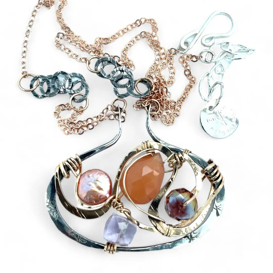 A darkened sterling silver pallete pendant necklace with bronze hammered swirls, featuring a series of faceted gemstones including amethyst and rhodocrosite, peach fresh water coin pearl, and crystal, complemented by a rose gold chain and a silver textured chain.