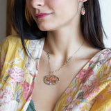 A darkened sterling silver pallete pendant necklace with bronze hammered swirls, featuring a series of faceted gemstones including amethyst and rhodocrosite, peach fresh water coin pearl, and crystal, complemented by a rose gold chain and a silver textured chain.