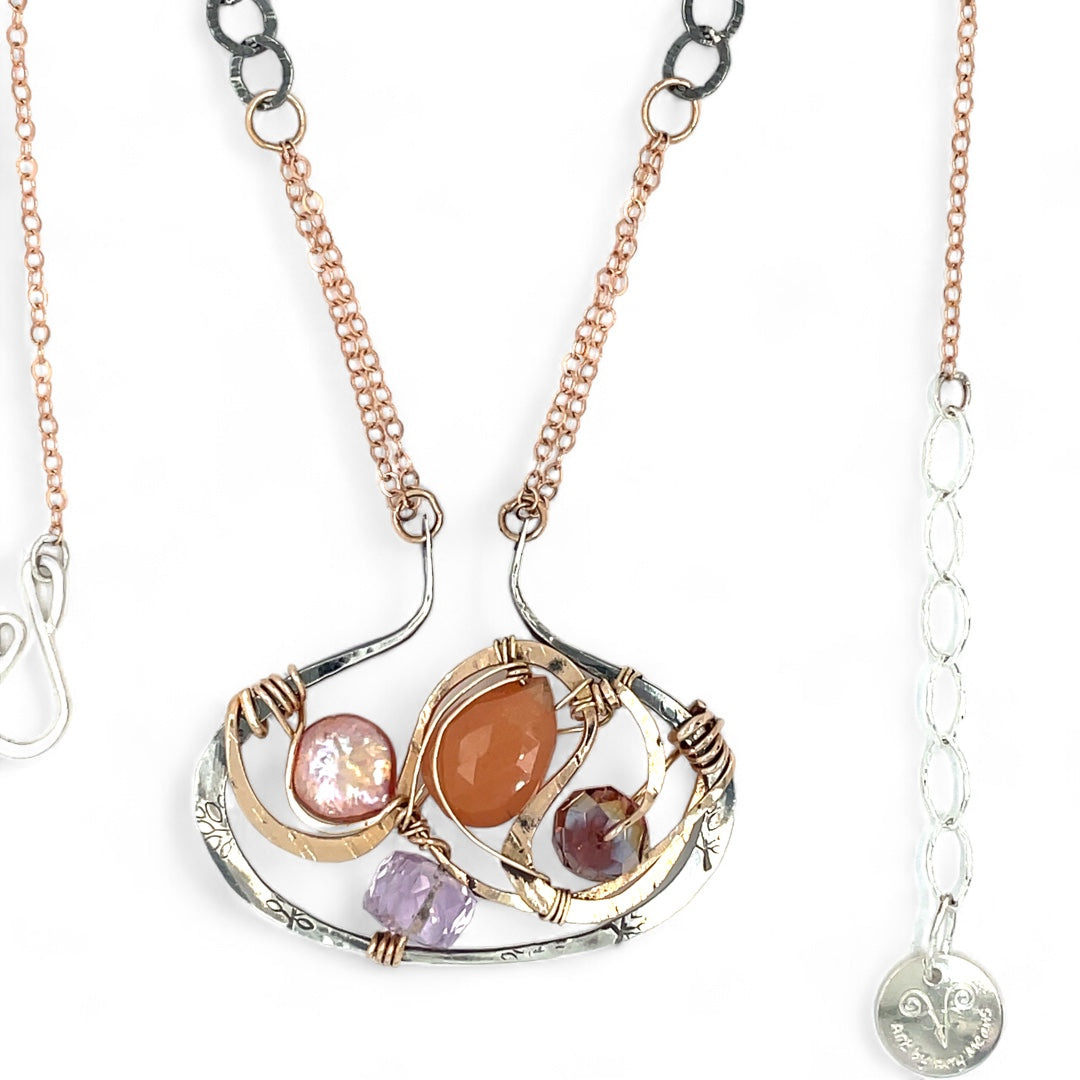 A darkened sterling silver pallete pendant necklace with bronze hammered swirls, featuring a series of faceted gemstones including amethyst and rhodocrosite, peach fresh water coin pearl, and crystal, complemented by a rose gold chain and a silver textured chain.