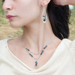 A pair of sterling silver leaf earrings with faceted green amethyst and blue tourmaline gemstones, wrapped in hand-formed hammered brass with sterling silver tube chain tops