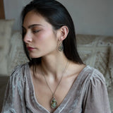 A pair of Prehnite smooth oval earrings with cool inclusions, adorned with hammered bronze, twisted oxidized sterling silver, and micro faceted watermelon tourmaline gemstones, attached to sterling silver French hooks.