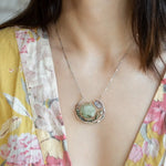 A Prehnite faceted coin gemstone with a mauve faceted moonstone set in bronze hammered wire inside a textured darkened sterling pendant, accented with green cat's eye faceted gemstones and a crystal finish.