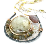 A Prehnite faceted coin gemstone with a mauve faceted moonstone set in bronze hammered wire inside a textured darkened sterling pendant, accented with green cat's eye faceted gemstones and a crystal finish.