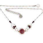 Red striped agate wrapped in hammered bronze wire suspended between sterling silver hammered petals holding faceted garnet and red crystal gemstones on a sterling silver bar chain necklace