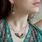 A person wearing a necklace with a hammered brass crescent pendant and double strands of sterling silver chains with Faceted cat's eye and black spinel gemstones and a clear quartz nugget