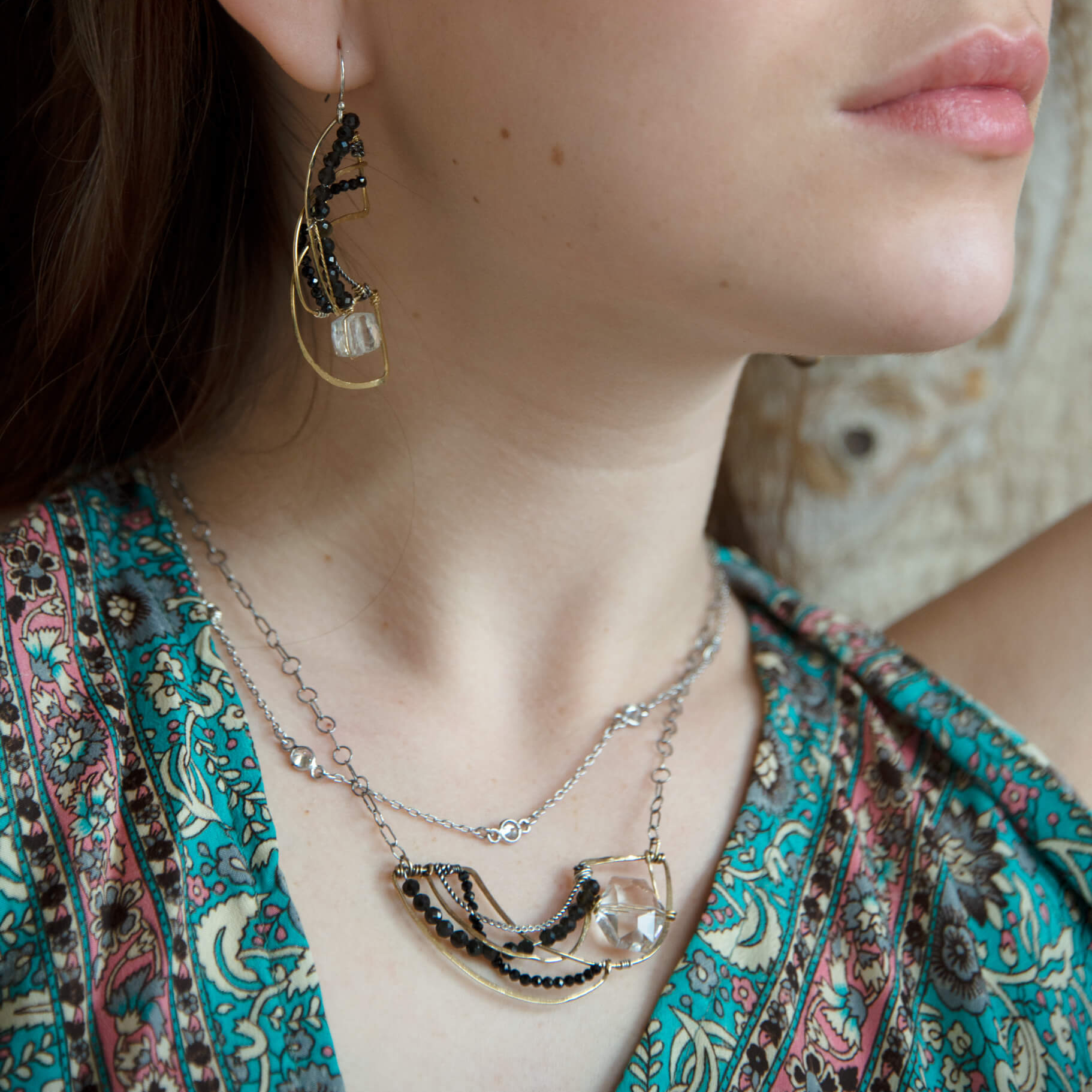 A person wearing a necklace with a hammered brass crescent pendant and double strands of sterling silver chains with Faceted cat's eye and black spinel gemstones and a clear quartz nugget