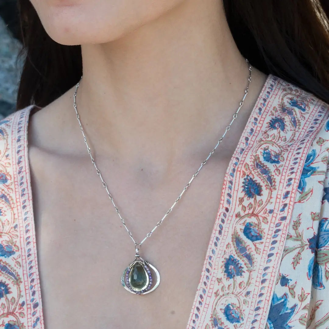 A necklace featuring a hand made thistle-shaped pendant with a green amethyst faceted drop gemstone and micro faceted lavender cubic zirconia gemstones, on a sterling silver bar chain.