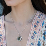 A necklace featuring a hand made thistle-shaped pendant with a green amethyst faceted drop gemstone and micro faceted lavender cubic zirconia gemstones, on a sterling silver bar chain.