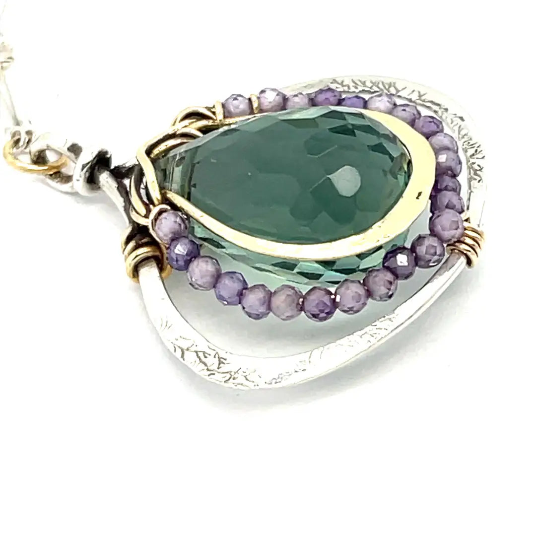 A necklace featuring a hand made thistle-shaped pendant with a green amethyst faceted drop gemstone and micro faceted lavender cubic zirconia gemstones, on a sterling silver bar chain.
