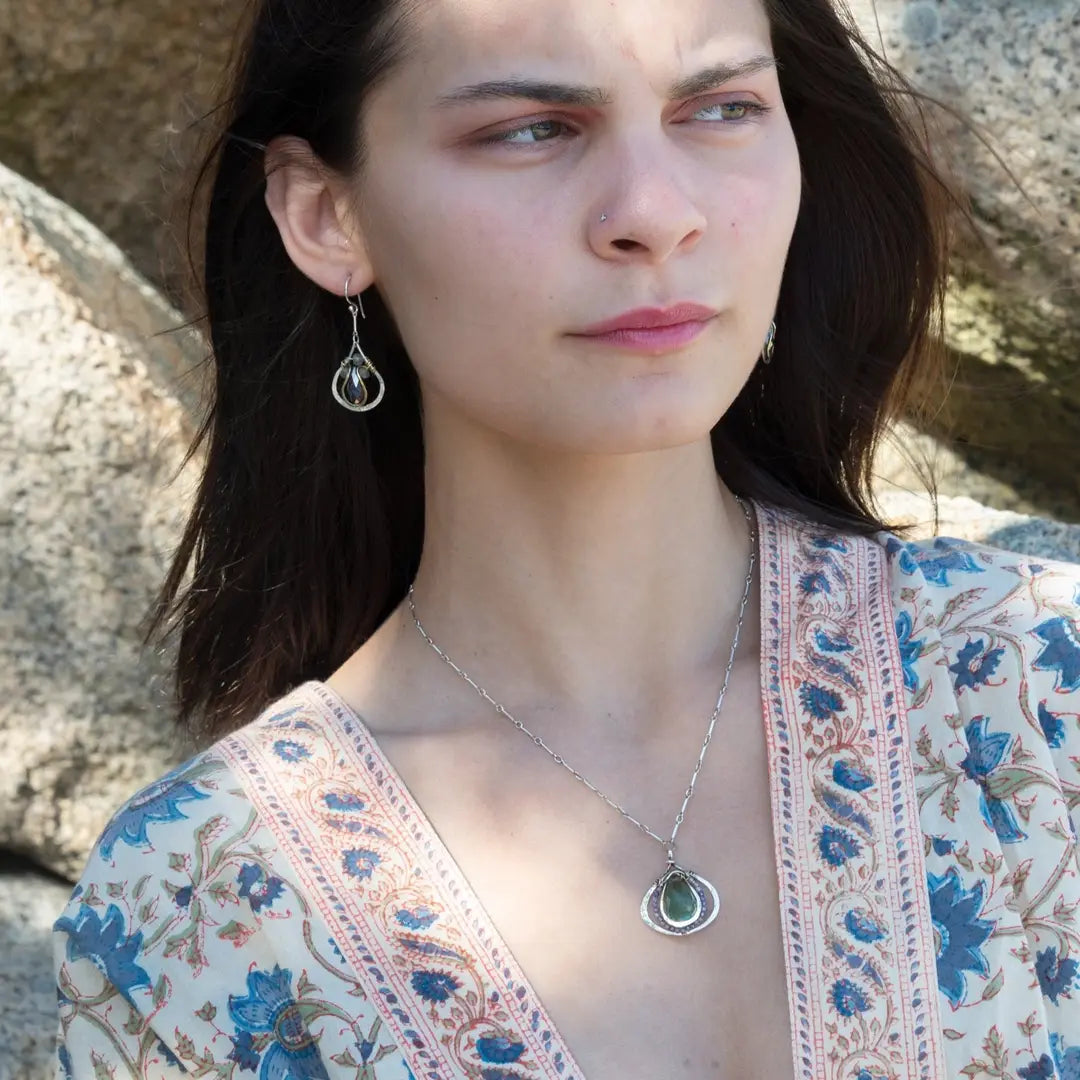 A necklace featuring a hand made thistle-shaped pendant with a green amethyst faceted drop gemstone and micro faceted lavender cubic zirconia gemstones, on a sterling silver bar chain.