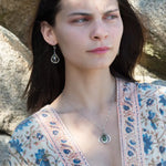 A necklace featuring a hand made thistle-shaped pendant with a green amethyst faceted drop gemstone and micro faceted lavender cubic zirconia gemstones, on a sterling silver bar chain.