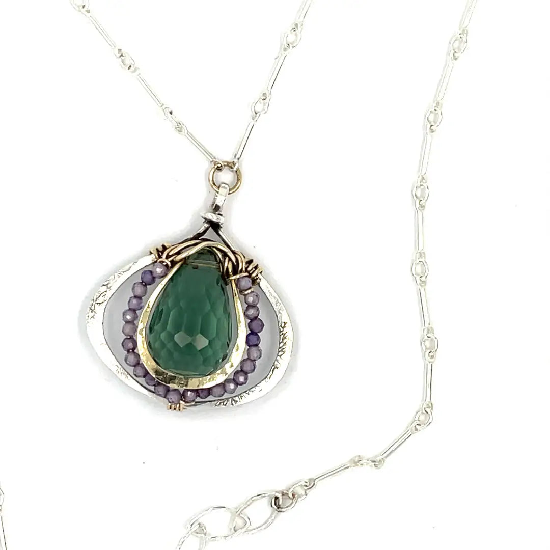 A necklace featuring a hand made thistle-shaped pendant with a green amethyst faceted drop gemstone and micro faceted lavender cubic zirconia gemstones, on a sterling silver bar chain.